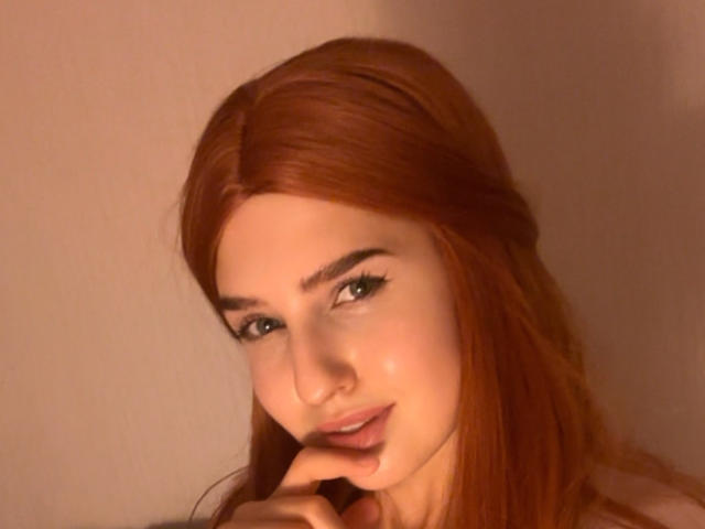 RosyAlma, con cabello rojo, tocándose el labio inferior con el dedo, tiene cejas oscuras, pestañas largas y una ligera sonrisa, mirando directamente a la cámara..