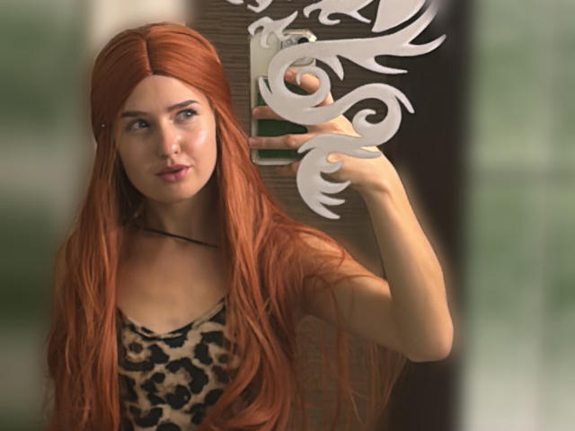 RosyAlma mit langen roten Haaren, die ein Top mit Leopardenmuster trägt, macht ein Selfie vor einem Spiegel, hält ein grünes Telefon und wirkt leicht gelangweilt, mit weißen, dekorativen Mustern im Spiegel..