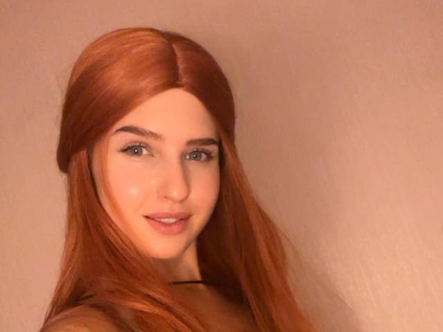 RosyAlma hat langes, rotes Haar, grüne Augen und ein süßes Lächeln mit dezentem Make-up. Ihre Brüste sind klein, und sie trägt einen schwarzen Choker. Sie schaut in die Kamera..