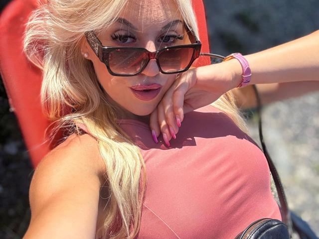CarolyneMarian-hot with blonde hair, wearing pink lipstick, large tits, pink nails, pink watch, and black sunglasses, leaning on red chair, sunlit, outdoors.