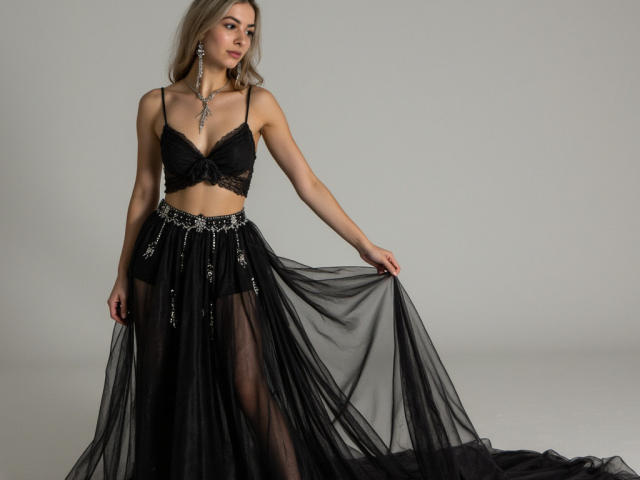 FancyRosie in black lace bra and sheer black skirt, holding flowing black fabric. She has blonde hair, a silver necklace, and a confident, sexy pose. Boobs visible.
