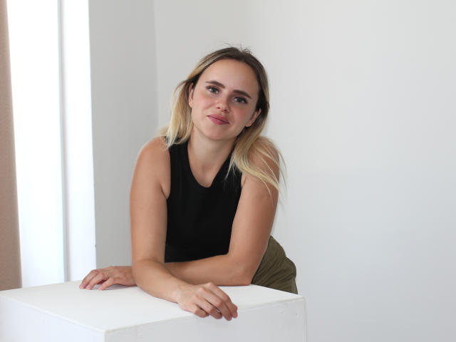 MilanaBloom with blonde hair, black sleeveless top, and olive green pants, leaning on a white table, smiling softly, hands resting on the table's edge.