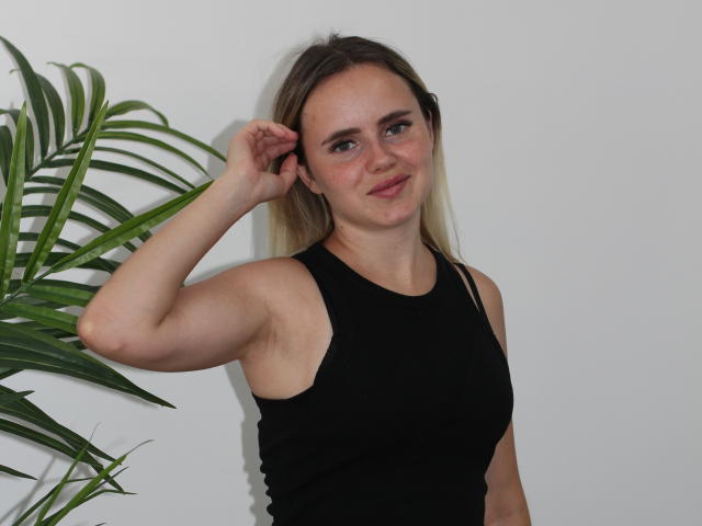 MilanaBloom with light brown hair, wearing a black sleeveless top, has her right arm raised, showing a relaxed smile, standing beside a green plant.
