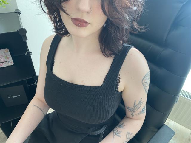 Khori with wavy brown hair, dark lipstick, nose ring, black tank top, tattoos on arms, sitting in black chair, showing her tits and small ass.