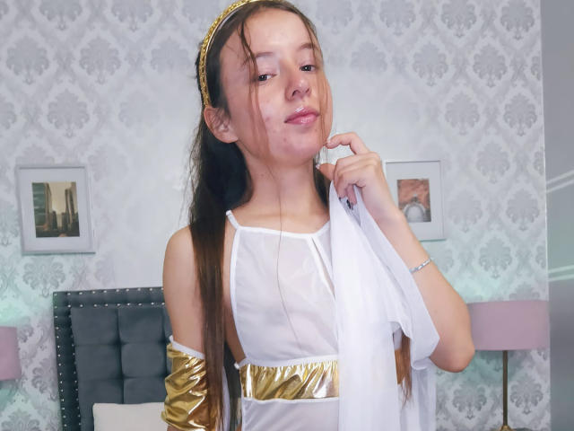 VioletaNova in a sheer white dress with gold arm bands and a gold headband, holding a white cloth near her mouth, showing her small tits and a cute smile.