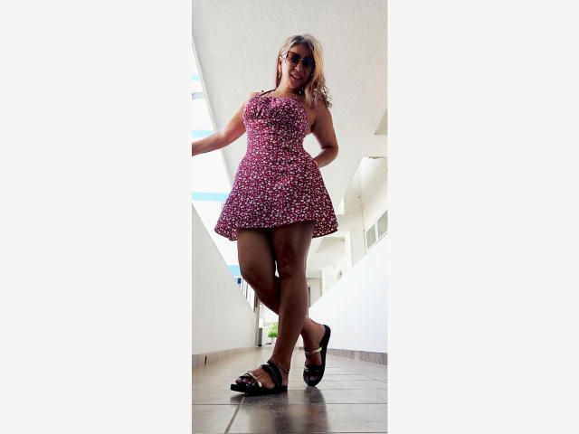 BrigiteWalsh with curly blonde hair, wearing a pink floral dress, black sandals, standing in a white hallway, looking down with a slight smile, hands on her hips.
