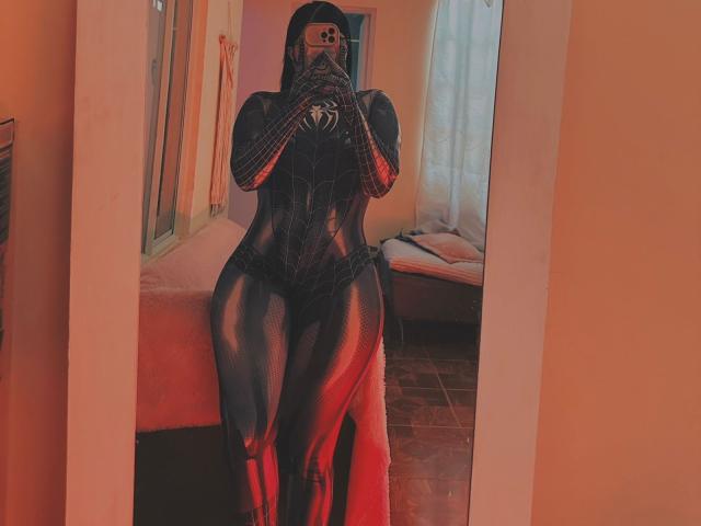 Mirror selfie of SweetYolandaX in a tight, shiny black Spider-Man suit, covering her face with a phone, sitting on a bed with red lighting, highlighting her curvy ass.