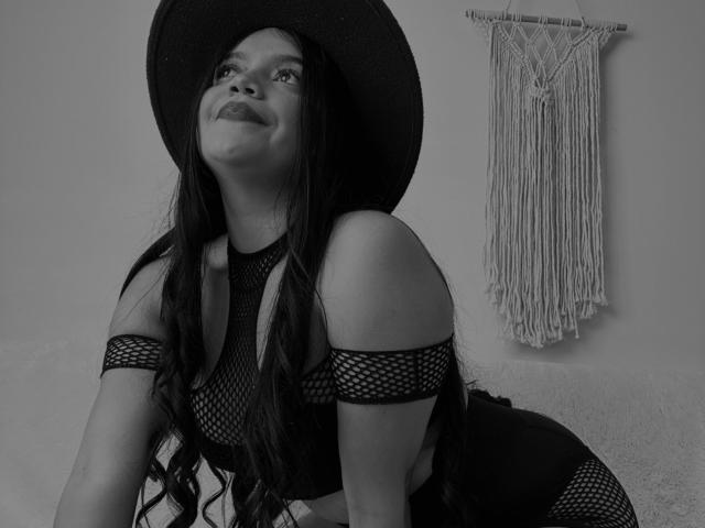 SweetYolandaX with long wavy hair, wearing a black mesh top and hat, looking up with a slight smile, her tits visible through the mesh, macramé wall hanging behind.