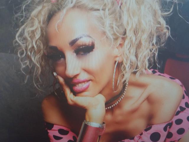 Barbie Ballerine - belle avec des cheveux blonds bouclés, un haut rose à pois, un trait d'eye-liner prononcé, des boucles d'oreilles cerceaux, un collier ras du cou, et une main sur la joue, avec une expression mignonne, des lèvres roses pulpeuses et une peau bronzée..