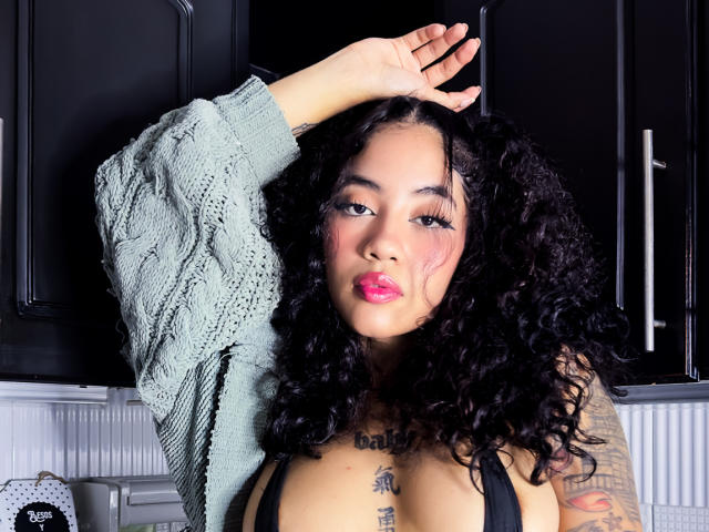 AvaRouge with curly black hair, pink lipstick, and tattoos, wearing a green sweater, arm raised, exposing black bra and large tits, standing in front of black cabinets.