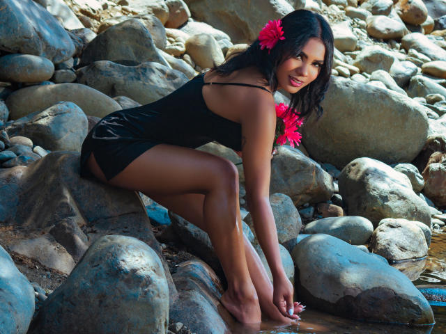 SusanFarrer with dark skin, black dress, pink flowers in hair, crouching on rocks, showing her ass, smiling at the camera, barefoot, sunlight, rocks in background.