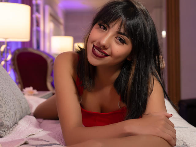KhloeWoods, with dark straight hair and red top, smiling at the camera, showing her tits, laying on a bed, arms crossed, in a cozy, lit-up room.