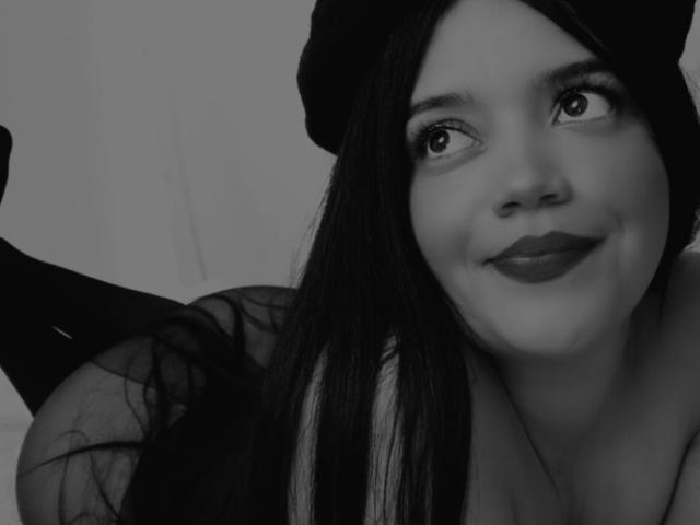 Black-and-white photo of SweetYolandaX with long dark hair, wearing a beanie, looking up with a smile, showing her cute face and wearing sheer black clothing.