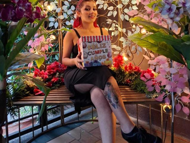 LizBonnet with red hair, black dress, showing book, holding cover, sitting on bench, legs crossed, showing thigh tattoo, surrounded by flowers, purple orchids, and pink blossoms.