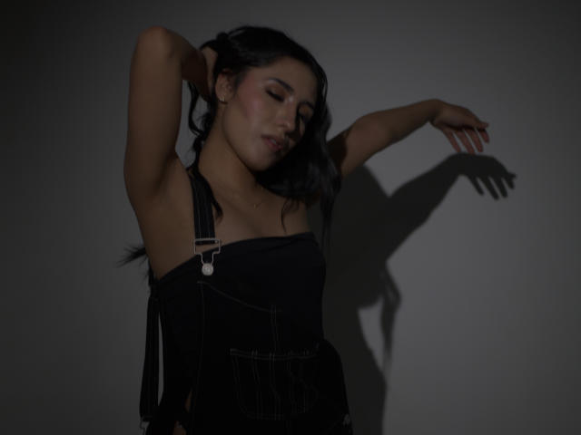 NickiClark has dark hair, eyes closed, wearing black suspenders, arms raised, shadow on wall, soft lighting, sensual expression, visible armpits, subtle makeup, relaxed posture, subtle tits.