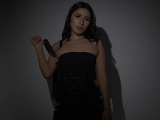 NickiClark with black wavy hair, wearing a strapless black dress, holding a knife in her right hand, looking slightly annoyed, dark shadow on her left side.