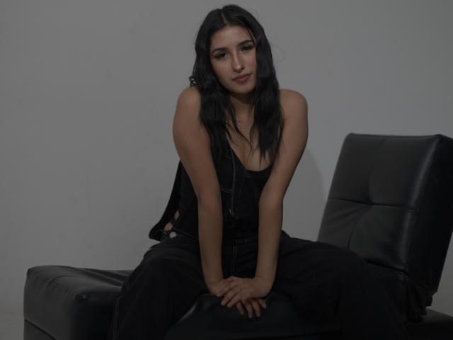 NickiClark with long black hair, sitting on black leather chair, wearing black dress, hands on chair, looking at camera, subtle smile, dark eyes, soft light, minimal shadow.