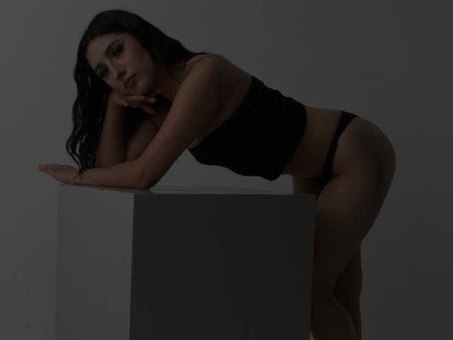 NickiClark with long black hair, leaning on a cube, wearing a black crop top and black panties, showing her ass and looking down with a hand on her cheek.
