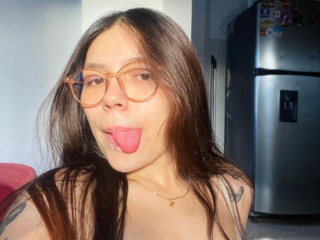 SophiaMarx has long brown hair, orange glasses, and is sticking out her pink tongue. She's wearing a gold necklace and has a tattoo on her left shoulder.