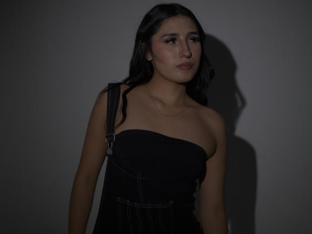 NickiClark with long dark hair, wearing a black one-shoulder dress, small tits, subtle makeup, shadow on gray wall, soft light, calm expression, simple necklace, dark background.