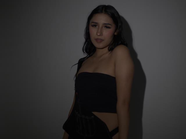 NickiClark has long black hair, wearing a black strapless top, exposing her tits and showing her ass with black pinstriped underwear, in dim lighting, looking slightly bored.