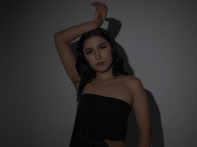 NickiClark with black wavy hair, dark strapless top, arm raised, shadow on wall, dark lighting, subtle makeup, confident expression, small gold necklace, slim build, medium tits.