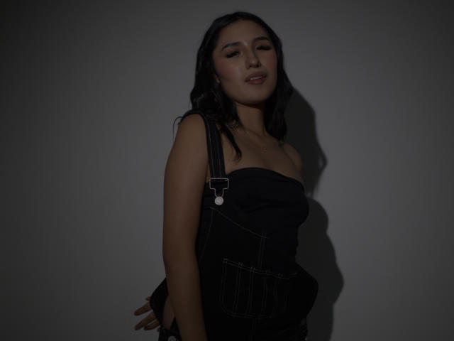 NickiClark with dark hair, wearing black strapless overalls, showing her tits slightly, arms behind back, shadowed, with eyes closed, looking relaxed and confident.