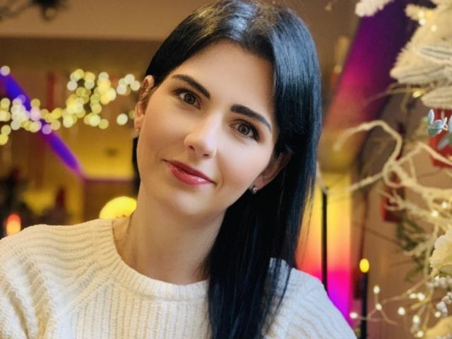 VioletaStyle has long black hair, a cute smile, and is wearing a white knitted sweater. She's looking at the camera with warm brown eyes and subtle makeup.