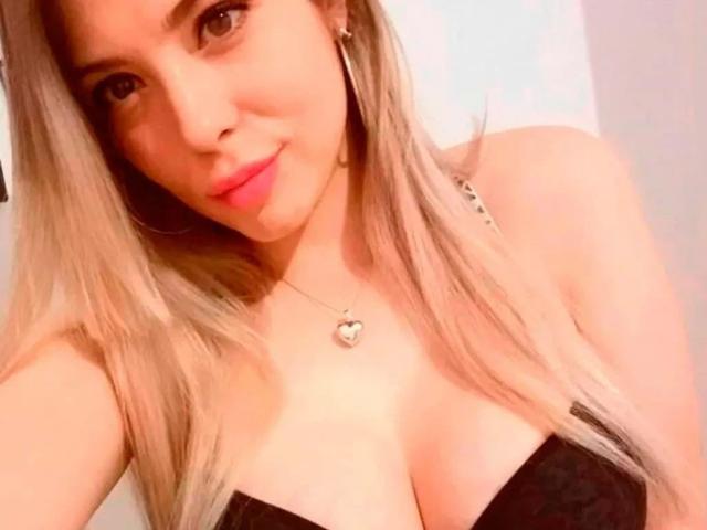 EvaBcn-hot with blonde hair, pink lipstick, hoop earrings, black strapless top showing her big tits, and a heart necklace, smiling at the camera.