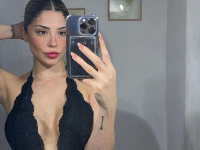 EvaBcn-hot with brown hair, red lipstick, and black lace halter top, holding a phone, showing her boobs, and a small tattoo on her right arm.