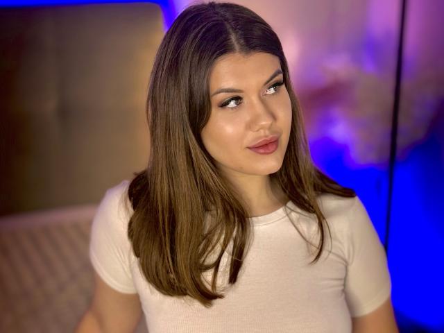 NikkiBust ha lunghi capelli castani, una maglietta bianca e un leggero sorriso con labbra rosate. I suoi occhi sono rivolti di lato e ha un trucco molto leggero..