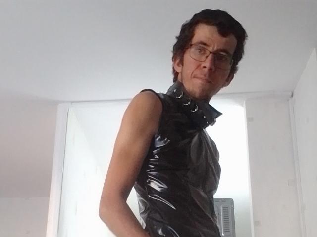 Bricebd with curly brown hair, glasses, and a black shiny sleeveless top with a high collar, standing in a white room, hands behind back, looking slightly confused.