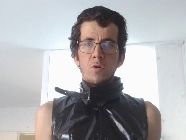 Bricebd with curly black hair, glasses, and a black shiny latex sleeveless top, looking slightly confused. His face has a bit of stubble. His butt is not visible.