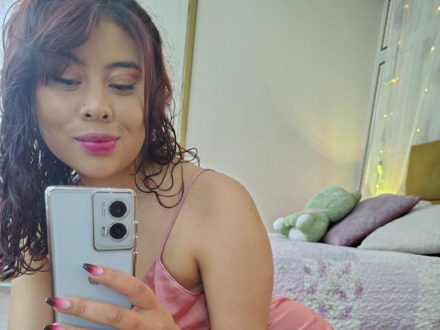 NoemiThornee with curly brown hair, pink lipstick, and pink nails, holding a phone, wearing a pink spaghetti-strap top, taking a selfie, showing her tits, in a bedroom.