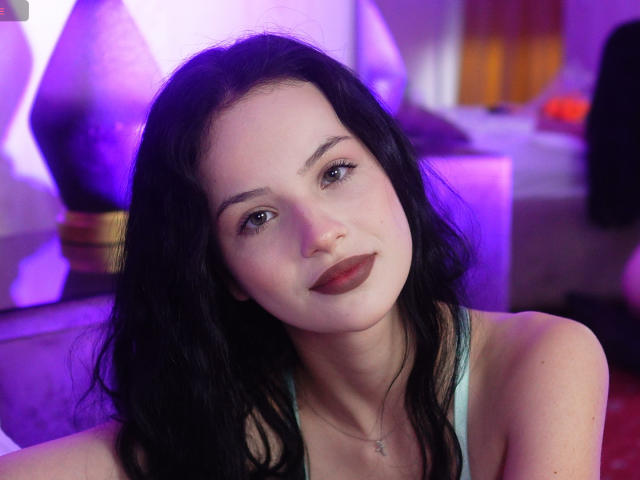 NatalieShaw with long black hair, pale skin, and red lipstick, wearing a green tank top, looking at the camera with a slight smile. Her tits are partially visible.