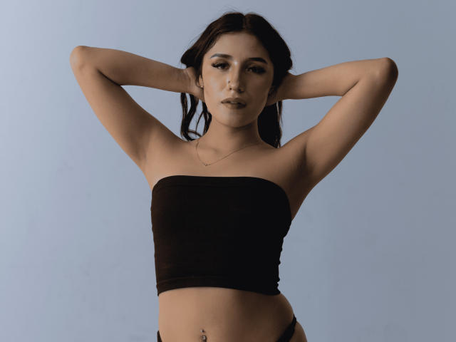 NickiClark with arms behind head, wearing a black strapless top, showing small tits, flat stomach, and a belly button piercing, against a plain background.