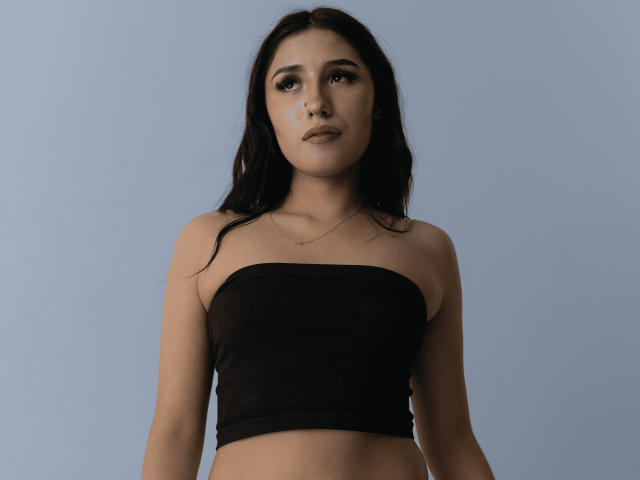 NickiClark has long black hair, a strapless black top, and a serious expression. She's standing against a blue background with a slight upward gaze. Boobs are covered.