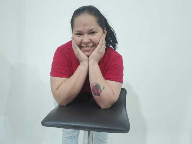 DulceHunny with black hair, red shirt, tattoo on right arm, smiling, hands on chin, leaning on black chair, white background, wearing light jeans, slightly chubby, happy expression.