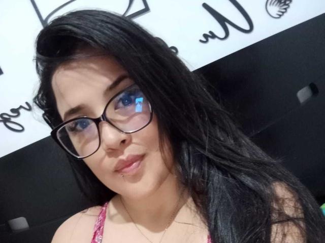 AlissonBeauty, con lunghi capelli neri, occhiali e una maglietta rosa, ha un piccolo piercing al labbro e occhi azzurri, mostrando un leggero sorriso. Il seno è visibile nella parte superiore dell'immagine..