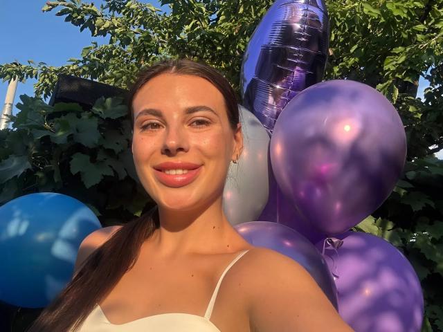 BlackFatal with brown poneytail, smiling, white spaghetti top, purple and blue balloons, shiny purple balloon, sunlit, green leaves, clear blue sky, soft glow on her face.