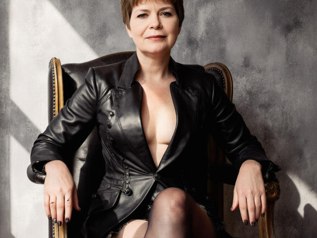 LoraMoon, short brown hair, black leather jacket, open to show small boobs, sitting on vintage chair, black stockings, confident smile, sunlight, concrete wall.