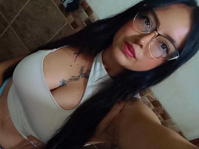 AilynRoss with long black hair, glasses, and pink lipstick, wearing a white halter top, showing her big tits and a tattoo on her right chest. 