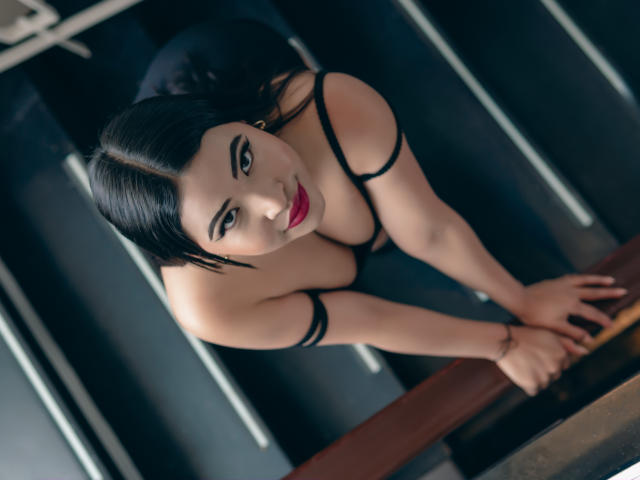 OliviGrace with black hair, red lipstick, and black lingerie, leaning forward with hands on a table, showing her tits and looking up with a seductive smile.