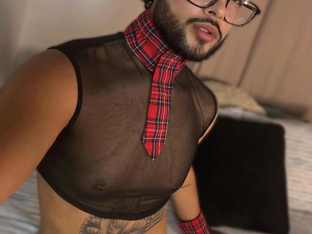 LeanPrensoom, wearing a sheer black top, red plaid tie, and glasses, with a beard, showing a tattooed lower belly and small nipples, in a bedroom.