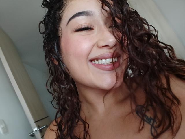 Lilith69Ember with curly brown hair, smiling with eyes closed, tongue out, showing teeth, and a tattoo on her chest. She has a nose ring and is topless.