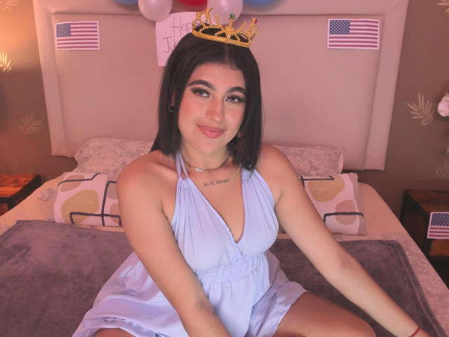 Daffne Addison, con un seno di media grandezza e un sorriso carino, è seduta su un letto indossando un abito azzurro chiaro e una corona dorata, con bandiere americane ai lati..