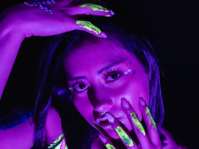 EmilysChoper's face with glowing neon green and purple paint on her hands, lips, and cheek, dark hair, black background, intense purple and green lighting.