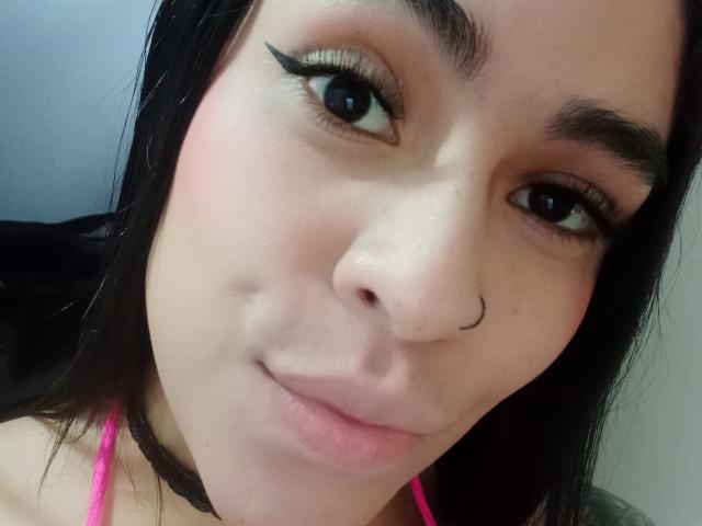 StellaBeckker's face with black hair, brown eyes, pink eyeliner, nose ring, pink lipstick, and slight smile. She's wearing a pink top, and has a black choker.