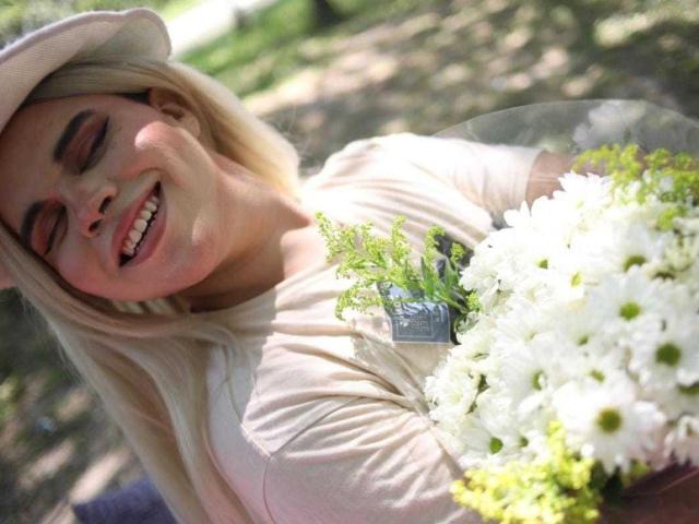 EvelyneRosse with blonde hair, smiling, wearing a beige hat, holding a bouquet of white daisies and greenery, eyes closed, sunlit, outdoors, casual beige shirt, happy, relaxed.