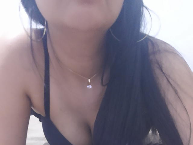 PatriciaLinda with long black hair, wearing a black bra that shows her tits, has a gold necklace with a small purple gem, and is leaning forward.